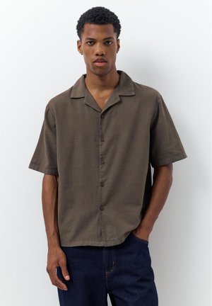 Mladý muž stojící před bílým pozadím, oblečený v volné hnědé košili s krátkými rukávy a tmavě modrých džínách.