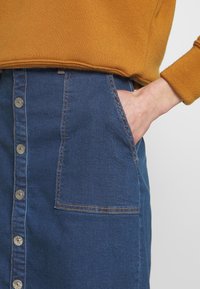 Džínová sukně s předním uzávěrem na knoflíky, tmavě modrá barva, postranní kapsy a oranžové švy. Spárována s hořčicovou halenou.