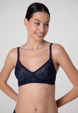Donna con capelli scuri sorride guardando di lato, indossando un reggiseno in pizzo blu navy con motivi floreali e bretelle sottili.