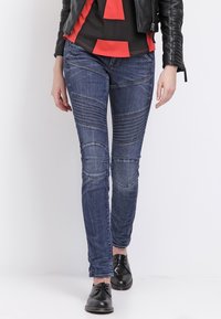 Jeans ajustados de color azul oscuro con detalle acolchado en las rodillas y rayas horizontales en los muslos, combinados con una chaqueta de cuero negra y zapatos.