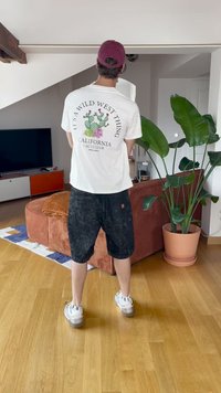 Bela majica s grafiko kaktusa, črne kratke hlače in bele športne copate. Stoji v moderni sobi z veliko zeleno rastlino in oranžno sedežno garnituro.