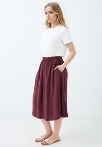 Jupe midi bordeaux avec taille élastique et poches latérales, accompagnée d'un t-shirt blanc ajusté à col rond et de sandales plates beiges.