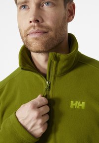 Zielona polarowa kurtka z wysokim kołnierzem, zamkiem błyskawicznym z przodu i logo na piersi. Miękka tekstura i dopasowany krój, z zapięciem na pół zamka.