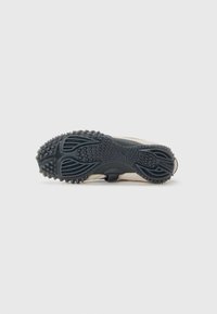 Beige sneaker laying on its side showing black rubber sole with textured grip and a small black loop near the heel.