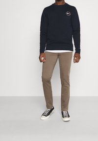 Felpa blu navy con logo, abbinata a pantaloni beige e scarpe nere. L'outfit presenta una vestibilità rilassata e una camicia bianca a strati sotto.