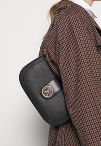 Sort struktureret håndtaske med en tekstureret overflade, guldfarvet hardware og et cirkulært logo. Båret over en ternet skjorte med en kædestrop.