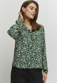 Grön blommig blus med svart bakgrund, med långa ärmar, krage och avslappnad passform. Blommönstret innehåller nyanser av grönt och vitt.