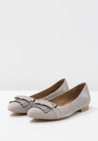 Un par de zapatos planos de ante gris claro con puntera redonda, lazos decorativos y flecos de metal en la parte frontal, exhibidos sobre una superficie blanca.