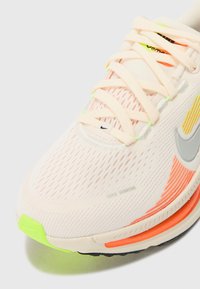 Zapatilla de running blanca de malla con acentos en durazno y gris, que presenta un upper texturizado, un talón verde neón y detalles reflectantes en los cordones.