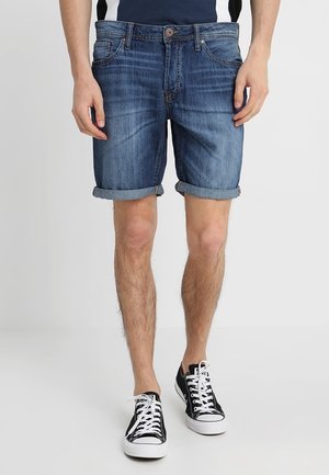 Shorts de mezclilla en azul medio con puños enrollados, cinco bolsillos y cierre frontal con botón. Llevados con zapatillas negras y blancas.