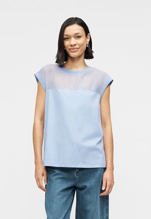 T-shirt bleu clair avec une partie supérieure transparente, des manches courtes retroussées et un texte brodé sur la poitrine. Associé à un jean bleu.