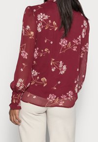 Blusa floreale bordeaux con maniche lunghe, tessuto trasparente, motivo floreale nei toni di rosa e beige, polsini arricciati e un design fluido.