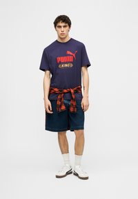 Νεαρός άνδρας που φοράει μπλε σκούρο t-shirt Puma King, τζιν σορτς, κόκκινο πουκάμισο με καρό δεμένο γύρω από τη μέση, λευκές κάλτσες και λευκά παπούτσια Puma, όρθιος.