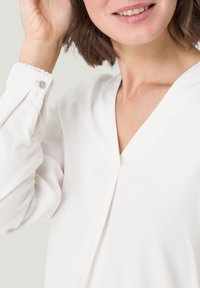 Femme en chemisier blanc avec des poignets festonnés, souriante et touchant ses cheveux courts et bruns.