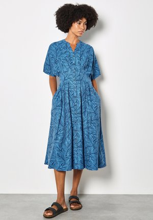 Kobieta z kręconymi włosami, ubrana w niebieską, midi sukienkę z krótkim rękawem, zapinaną na guziki z wzorem liści i czarnymi sandałami, stojąca na tle białej ściany.