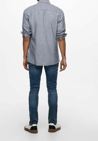 Camisa de botones gris con mangas enrolladas, combinada con jeans ajustados azules y zapatillas blancas con acentos negros. Atuendo simple y casual.
