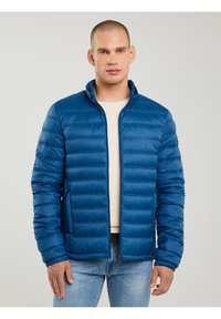 Rövid hajú férfi, kék steppelt dzsekit visel bézs pulóver fölött és világoskék farmert, egyszerű háttér előtt áll.