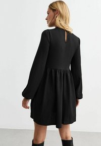 Vestido negro texturizado con mangas largas y escote redondo, que presenta un diseño de espalda con escote en forma de lágrima y falda acampanada. Ideal para uso casual.