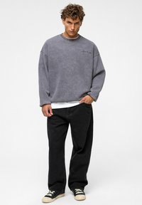 Grijze oversized sweatshirt met ronde hals, textuurstof en logodetail; gecombineerd met zwarte losse jeans en zwarte sneakers.