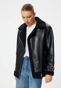 Svart skinnjacka med fårskinnskrage och manschetter, asymmetrisk dragkedjestängning och bälte-detalj. Bärs över en ljus skjorta och jeans.