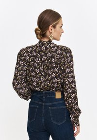 Kobieta z włosami upiętymi w bun, w złotych kwiatowych kolczykach, długim czarnym blouse z różowymi i żółtymi kwiatami oraz ciemnoniebieskich dżinsach o wysokim stanie.