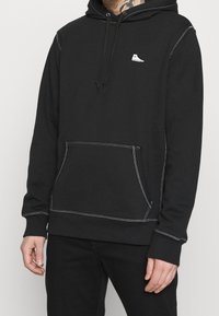 Felpa nera in misto cotone, con tasca frontale, cuciture bianche a contrasto, cordini regolabili e un piccolo logo sul petto.