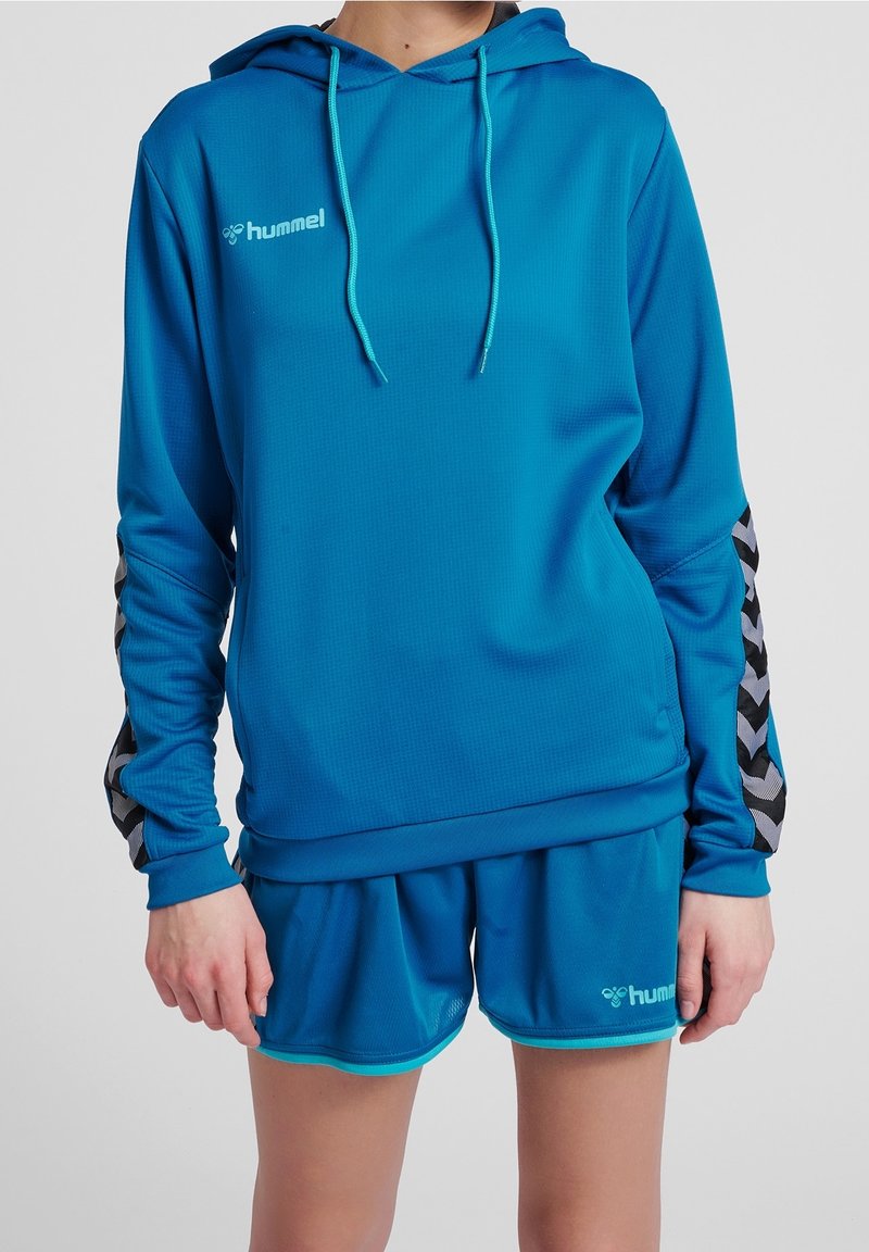 Sweat à capuche turquoise en tissu texturé, avec une capuche et des manches à motif chevron gris. Associé à un short assorti, également en turquoise.