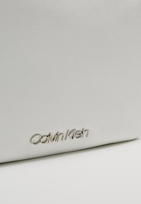 Biała torebka z teksturowanej syntetycznej skóry z metalowym logo "Calvin Klein" w kursywie z przodu. Prosty design bez dodatkowych akcentów.