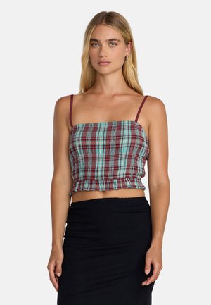 Femme blonde portant un haut court à carreaux avec de fines bretelles bordeaux et une jupe noire taille haute, debout devant un fond blanc.