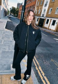 Fekete steppelt bomber dzseki arany cipzárral és nap emblemmel, fekete, szárában grafikus részleteket viselő trapéz nadrággal párosítva.