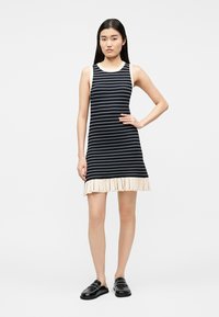 Junge Frau trägt ein ärmelloses, schwarz-weiß gestreiftes Kleid mit einem cremefarbenen, gerafften Saum und schwarzen Slippern.