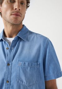 Camisa de mezclilla de manga corta en azul claro con botones. Presenta un bolsillo de parche y un bordado sutil en el pecho. Textura suave y corte relajado.