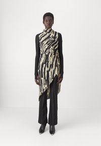 Robe asymétrique noire et crème avec motif abstrait, design drapé, associée à un pantalon noir ajusté et des talons noirs pointus.