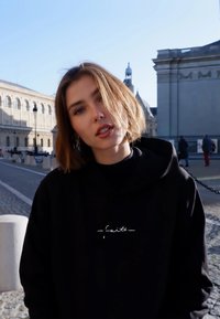 Felpa nera con cappuccio caratterizzata da un ricamo bianco con la parola “faith”; tessuto morbido, vestibilità rilassata, cuciture visibili, indossata all'aperto su uno sfondo soleggiato.