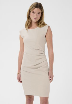 Žena so svetlohnedými vlasmi, oblečená v bezrukávových béžových šatách po kolená, stojaca pred jednoduchým bielym pozadím.