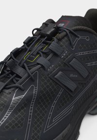 Gros plan sur une chaussure de sport noire avec un mesh texturé, des détails réfléchissants, des lacets élastiques et un mécanisme de verrouillage des lacets sur la languette.