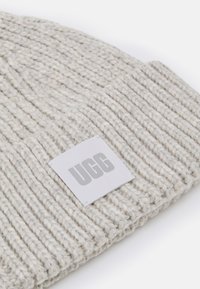 Lys grå strikket lue med ribbestruktur og et rektangulært UGG-logo-patch sydd på forsiden, med et mykt, tykt garn-design.