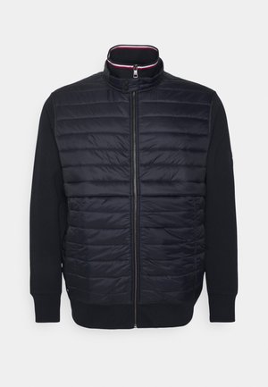 Czarna pikowana kurtka z przodu z pełnym zamkiem, zapięciem na zatrzask kołnierza oraz jednolitymi czarnymi rękawami i prążkowanymi mankietami.