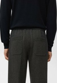 Pantaloni de trening gri închis cu finisaj texturat, talie elastică și două buzunare în spate. Purtat cu o cămașă neagră cu mâneci lungi.