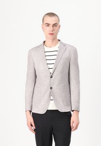 Blazer gris clair en tissu texturé, à fermeture à un bouton, avec deux poches avant. Porté par-dessus un pull blanc à rayures et un pantalon sombre.