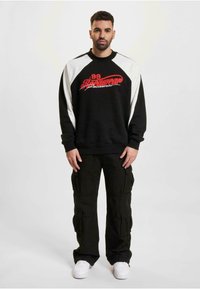 Sweatshirt noir avec texte graphique rouge et manches raglan blanches, associé à un pantalon cargo noir avec de multiples poches et des baskets blanches.