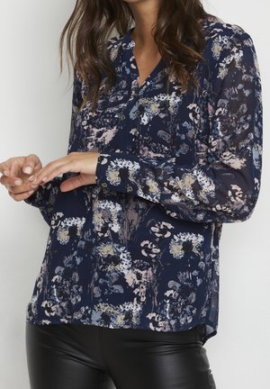 Kvinde iført marineblå, gennemsigtig bluse med blomsterprint og lange ærmer samt sorte læderbukser, hænderne let sammenføjede foran.