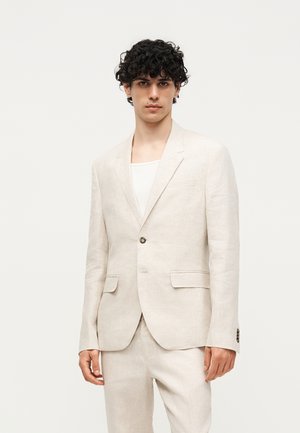 Junger Mann mit lockigem dunklem Haar, der eine hellbeige Anzugjacke und passende Hose über einem weißen Hemd trägt, steht vor einem einfarbigen Hintergrund.