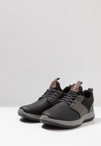 Zapatos deportivos negros con un exterior suave, acentos grises, perforaciones transpirables y detalles de cuero marrón cerca de los cordones. Suela de goma.