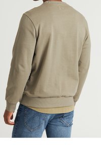 Felpa beige con finitura testurizzata e polsini a coste, vista da dietro. Abbinata a jeans blu, mostra uno stile casual.