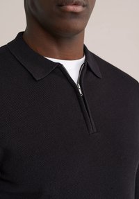 Črni pleteni pulover z zadrgo na ovratniku, s teksturirano tkanino in belo notranjo plastjo, ki je vidna ob izrezu vratu.