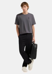 T-shirt grigio a maniche corte con logo, abbinato a pantaloni neri su misura e sneakers bianche, mentre tiene una borsa per laptop nera.