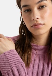 Light pink ribbed knit sweater with a round neckline; model's hand resting on the sleeve. Close-up focus on facial features and hair.