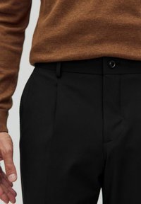 Zwarte op maat gemaakte broek met een platte voorkant, voorzien van een enkele knoopsluiting en zijzakken. Draagt een bruin sweatshirt.