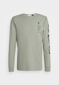 Hellgrünes langärmeliges T-Shirt aus Baumwolle. Es zeigt schwarzen Text auf der Brust und grafische Akzente entlang des linken Ärmels.